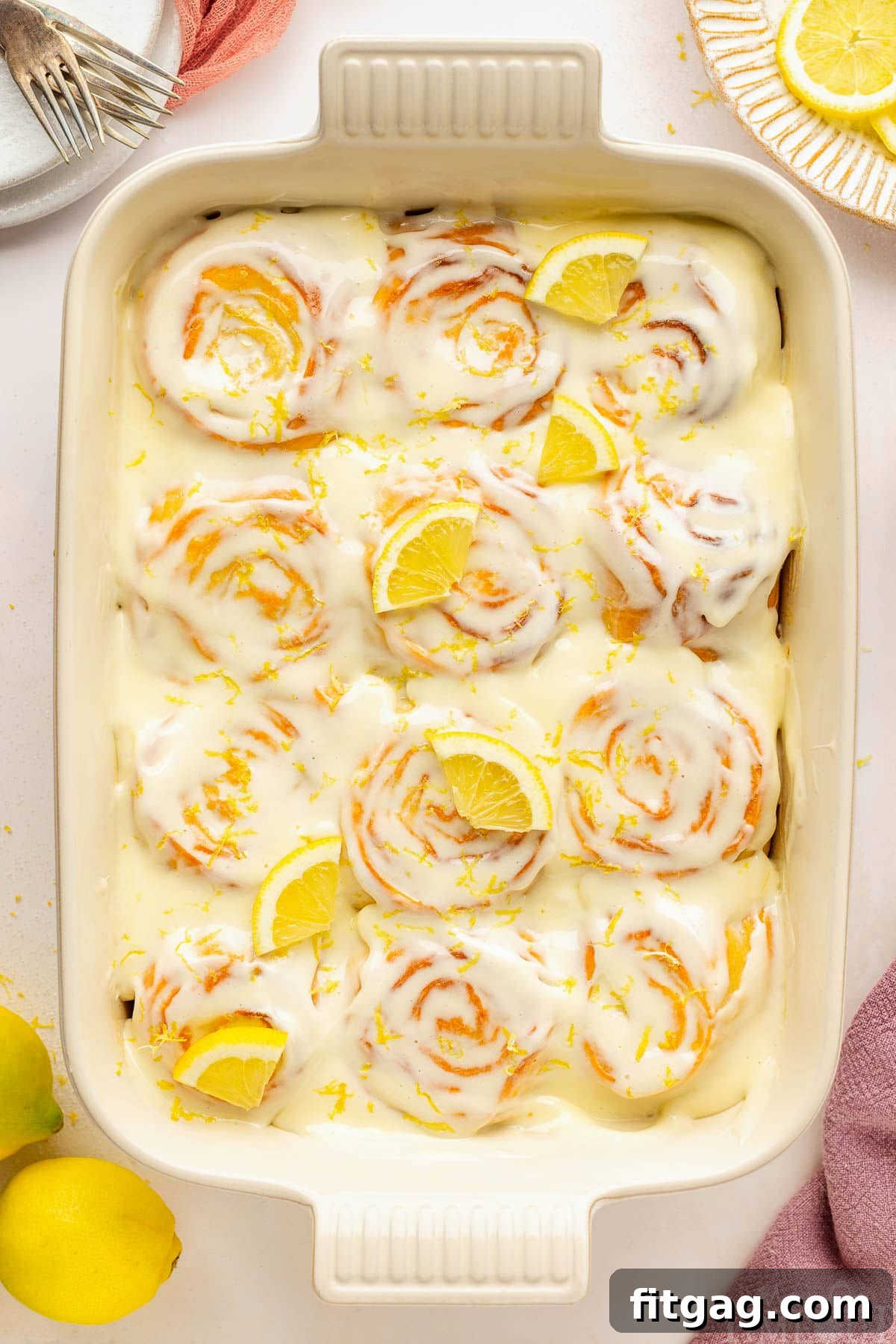 Sunshine Lemon Rolls 2 A top-down view of a pan filled with freshly baked lemon sweet rolls, generously drizzled with a creamy lemon glaze and adorned with fresh lemon slices and zest, set on a clean white surface.
