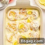 An overhead view of a pan of lemon sweet rolls topped with a lemon cream cheese glaze, lemon slices and lemon zest. Baking dish is white.