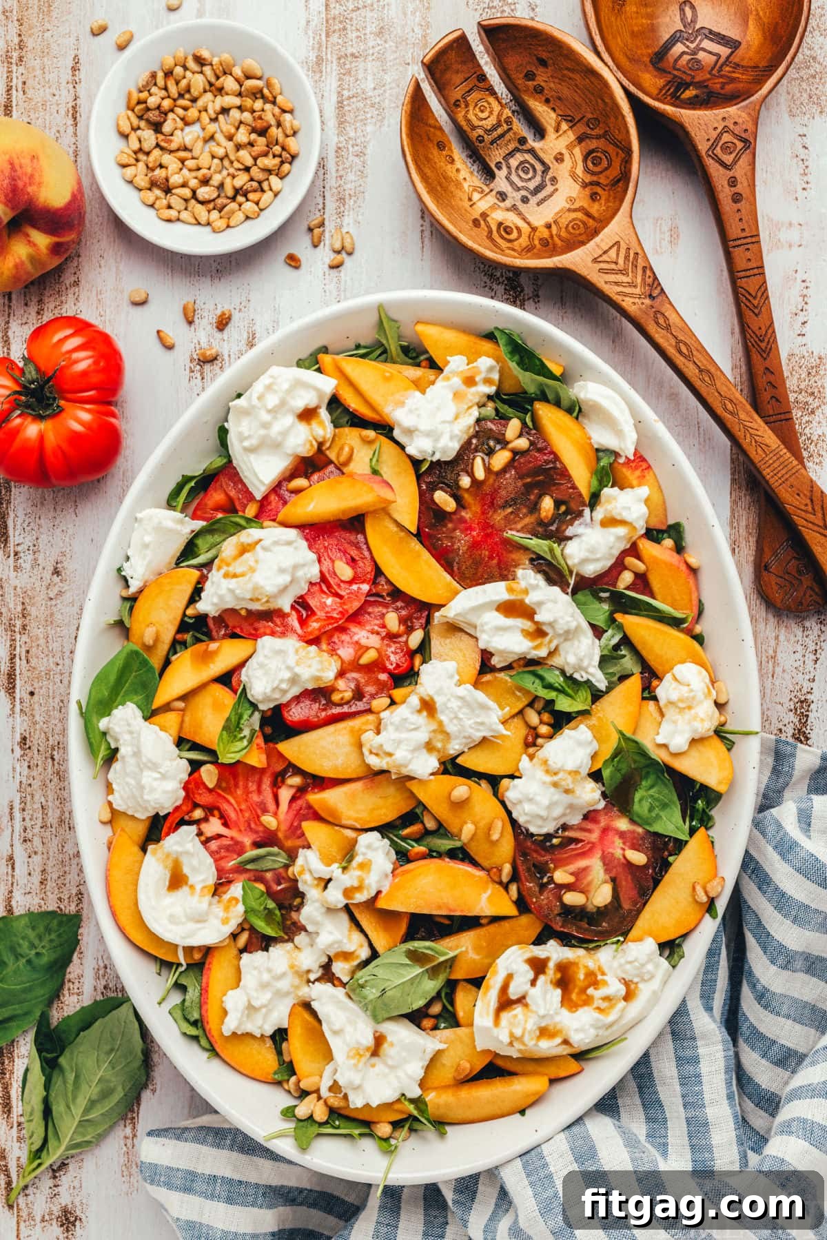 Fresh peach salad made with burrata and heirloom tomatoes, all topped with a balsamic vinaigrette. The salad is served on a white oval platter and garnished with toasted pine nuts and fresh basil.