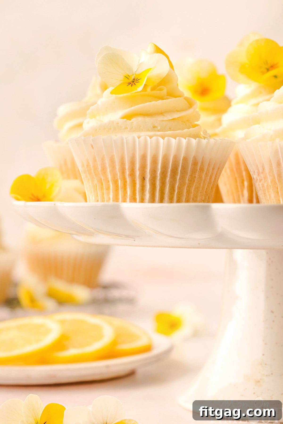An array of freshly baked and frosted lemon cupcakes arranged on a white scalloped cake stand, with more cupcakes and lemon slices visible in the background.