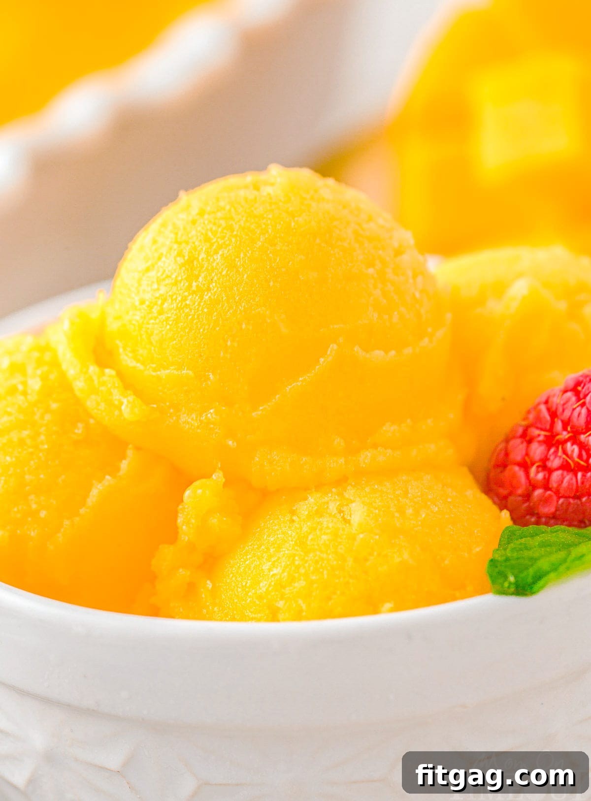 Scoops of vibrant yellow mango sorbet in a pristine white bowl, garnished with a fresh mint leaf, set against a bright, summery background.