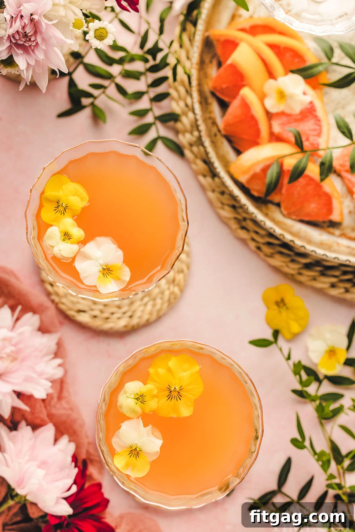 A top-down view of two French Blonde Cocktails in delicate coupe glasses, beautifully presented on a pink surface with citrus slices and delicate edible flower garnishes.