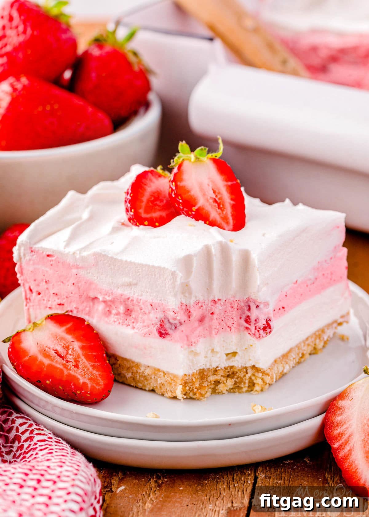A perfectly cut square of no-bake strawberry lasagna on a round white plate, generously topped with whipped cream and fresh strawberry slices. A bowl of whole, ripe strawberries is blurred in the background, suggesting the fresh ingredients used.