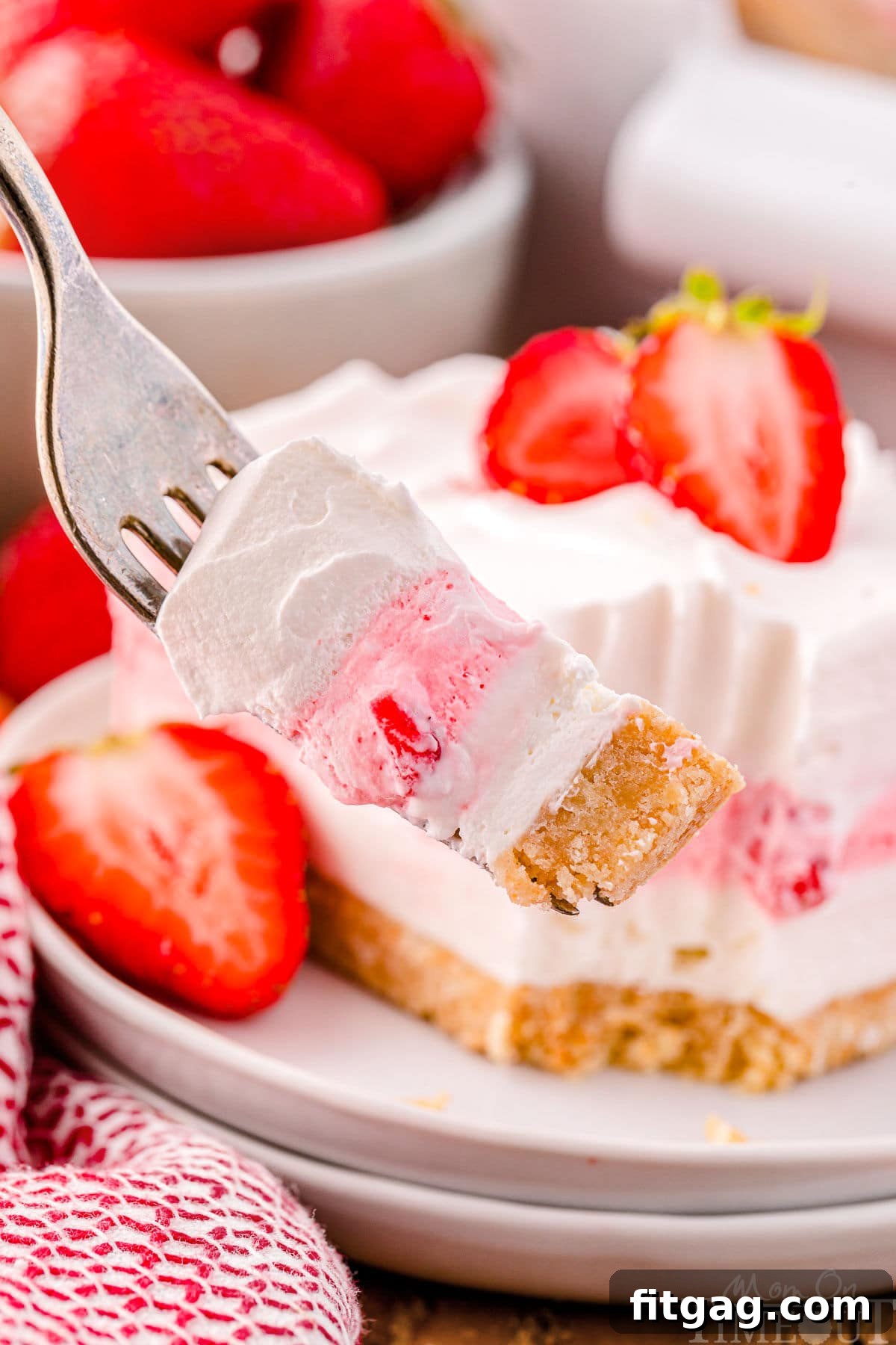 A fork lifts a perfect bite of Strawberry Lasagna, showcasing a clear view of its distinct layers: the crunchy cookie crust, the creamy cheesecake, and the vibrant strawberry gelatin, topped with fluffy whipped cream and a piece of fresh strawberry.