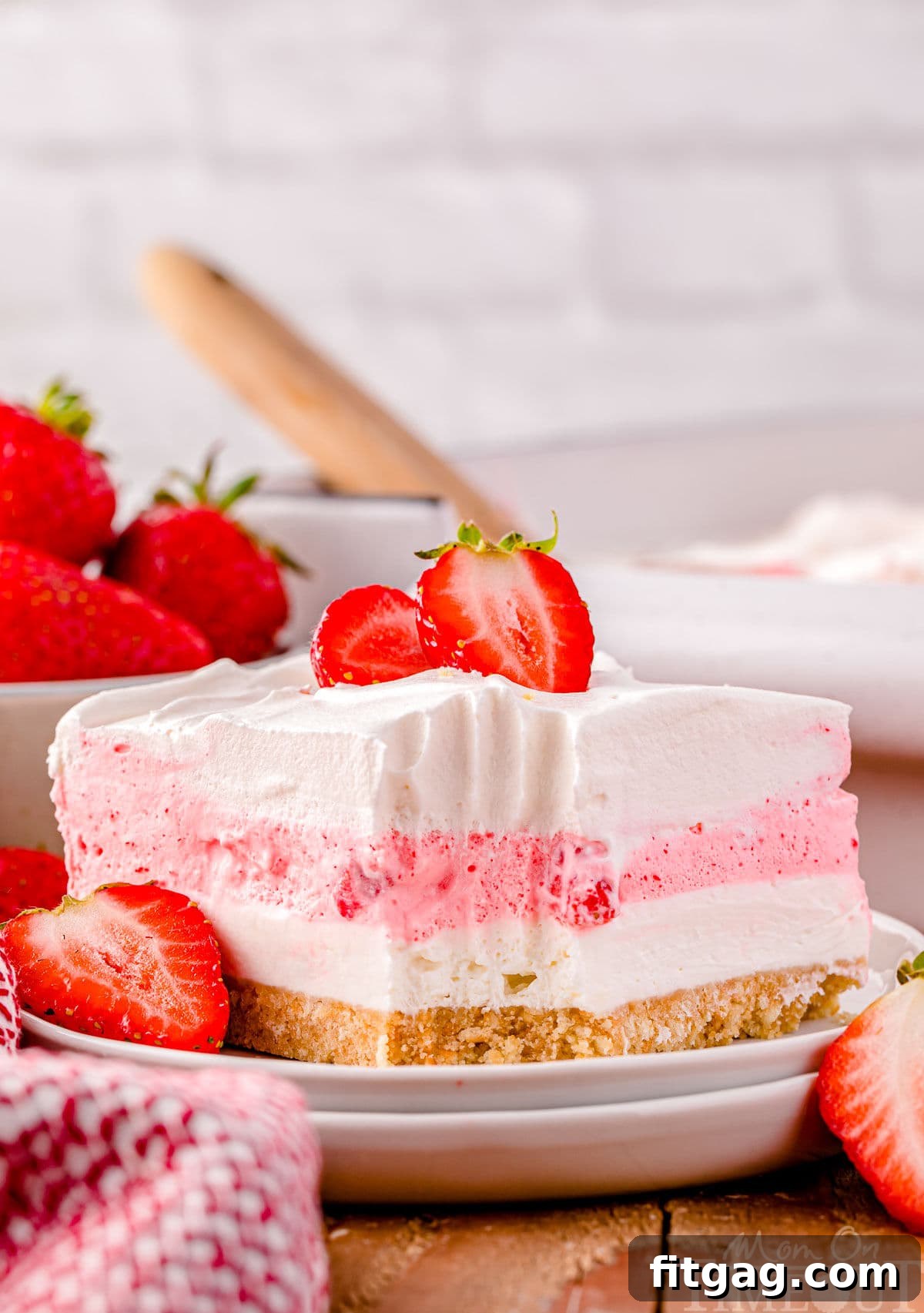A perfectly portioned slice of No-Bake Strawberry Lasagna is showcased on a white dessert plate, with the distinct layers clearly visible. A delicate sprig of fresh mint and a few extra strawberry slices garnish the plate, emphasizing the dessert's fresh ingredients.