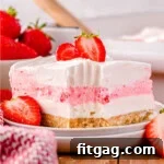A slice of strawberry lasagna on a white plate topped with fresh strawberries. The baking dish with remaining dessert and a bowl of fresh strawberries are visible in the background.