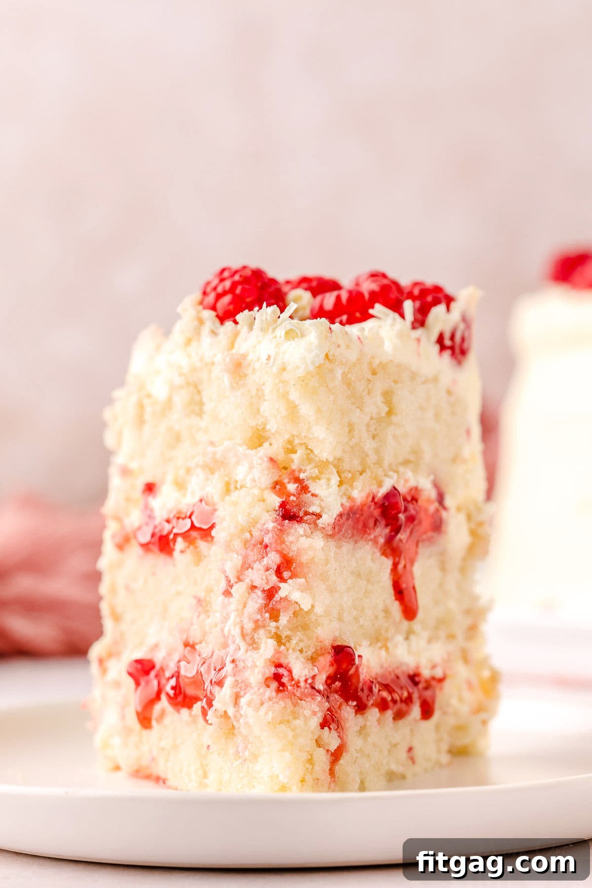 Slice of white chocolate raspberry cake on a white round plate topped with fresh raspberries and white chocolate curls.
