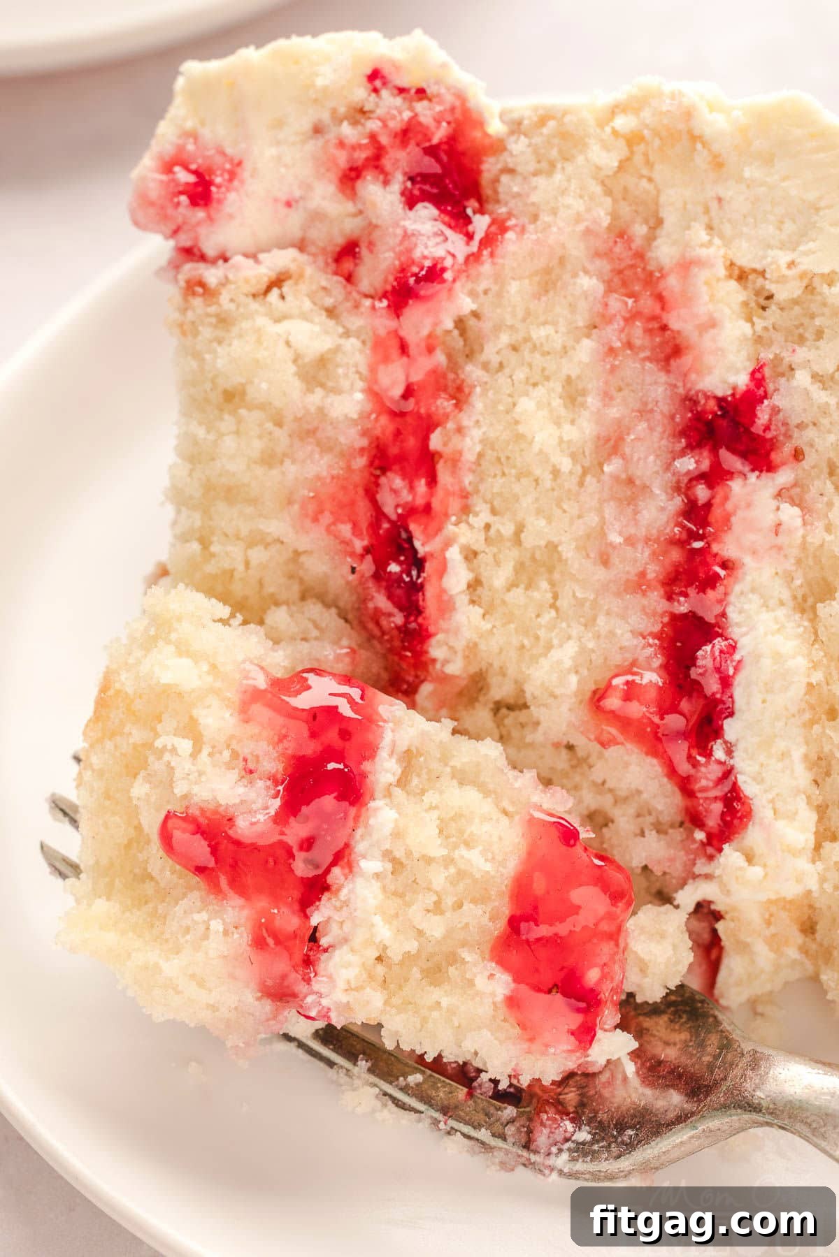 slice of raspberry white chocolate cake on white round plate. Fork has cut off a bite of the cake.