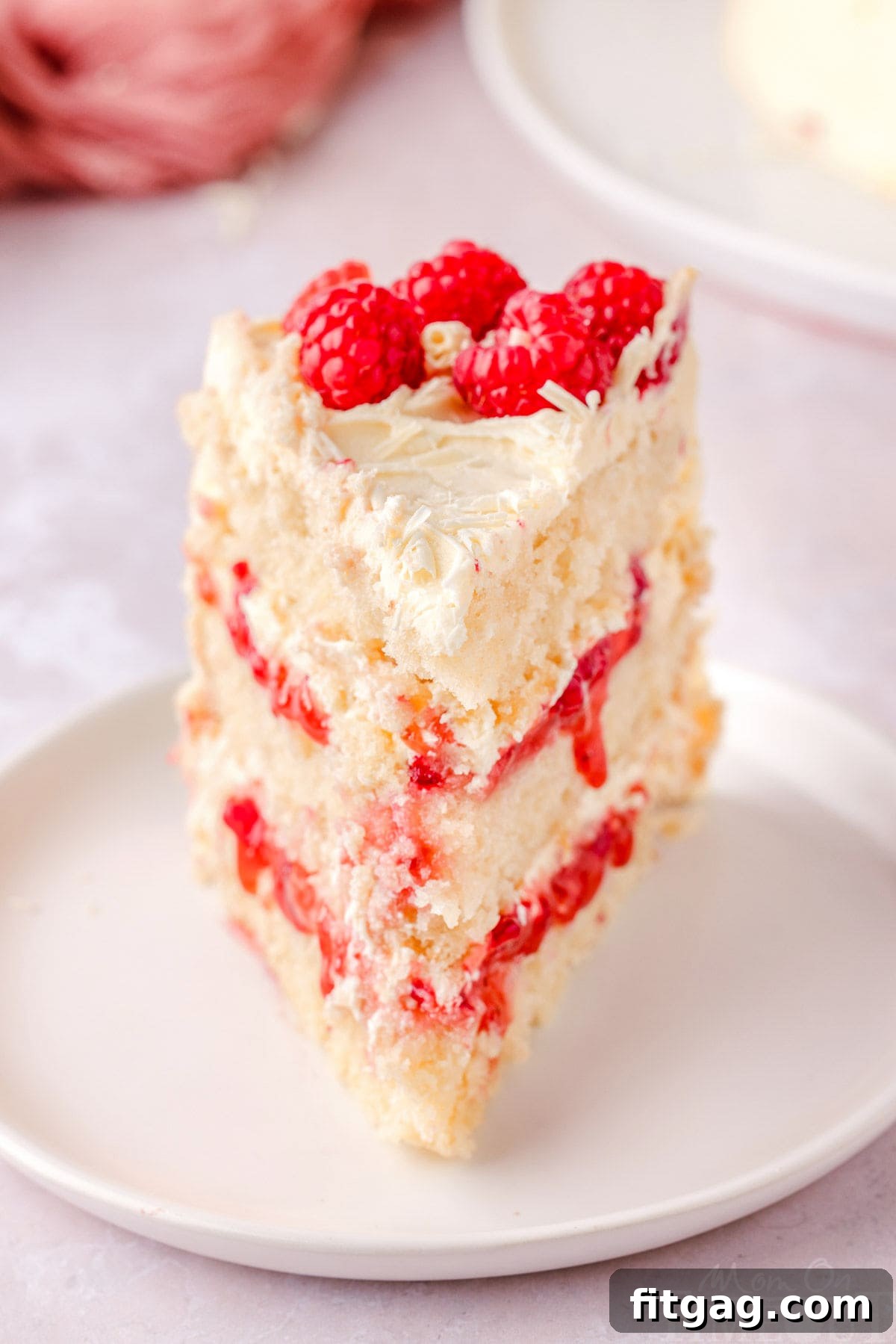 Slice of white chocolate raspberry cake on a white round plate topped with fresh raspberries and white chocolate curls.