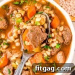 A top down view of a spoon in a bowl of beef and barley soup.