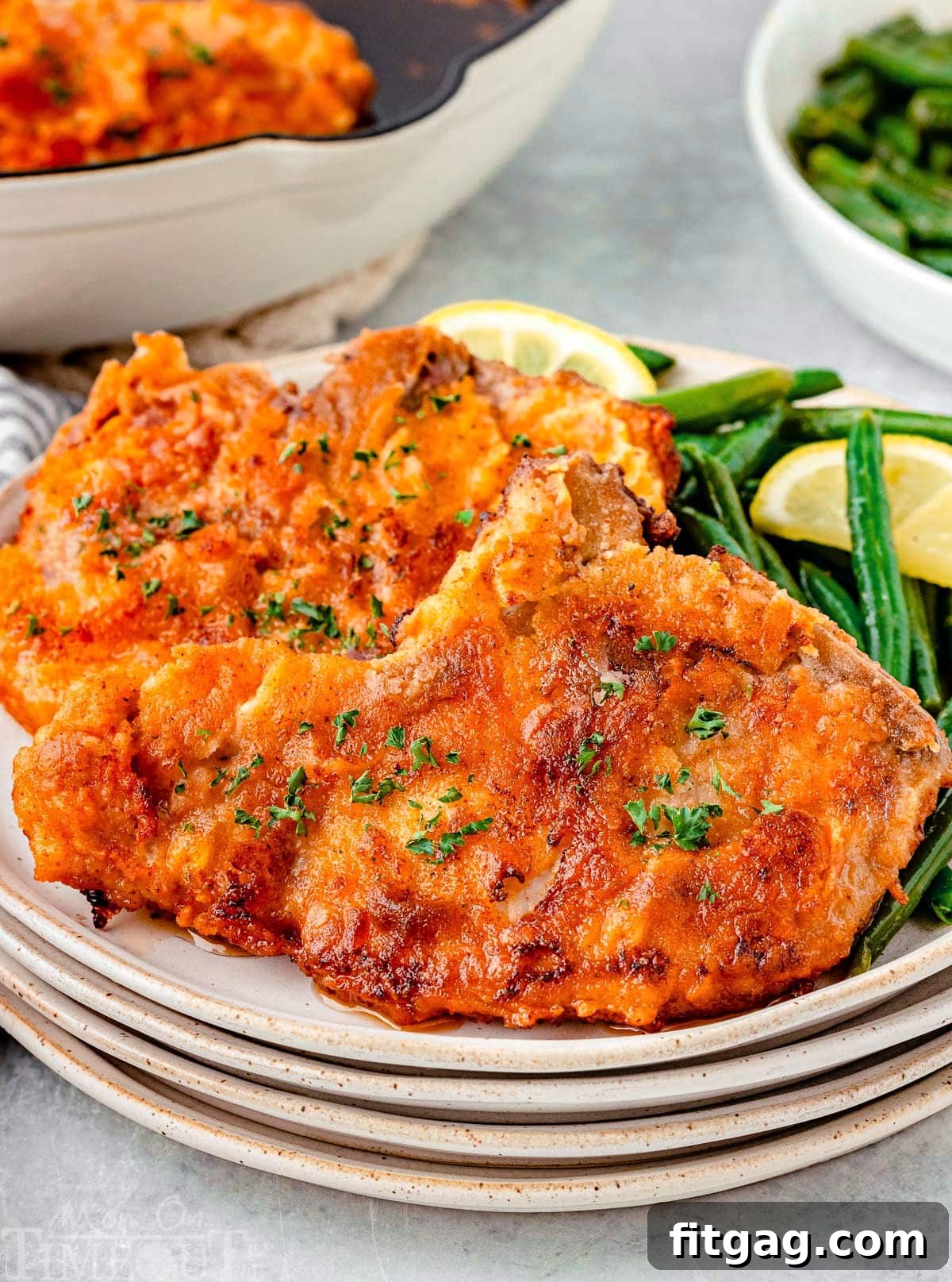Golden-Crusted Pork Steaks 2 Two fried pork chops on a stack of small round plates. Pork chops are plated with green beans and a lemon wedge.