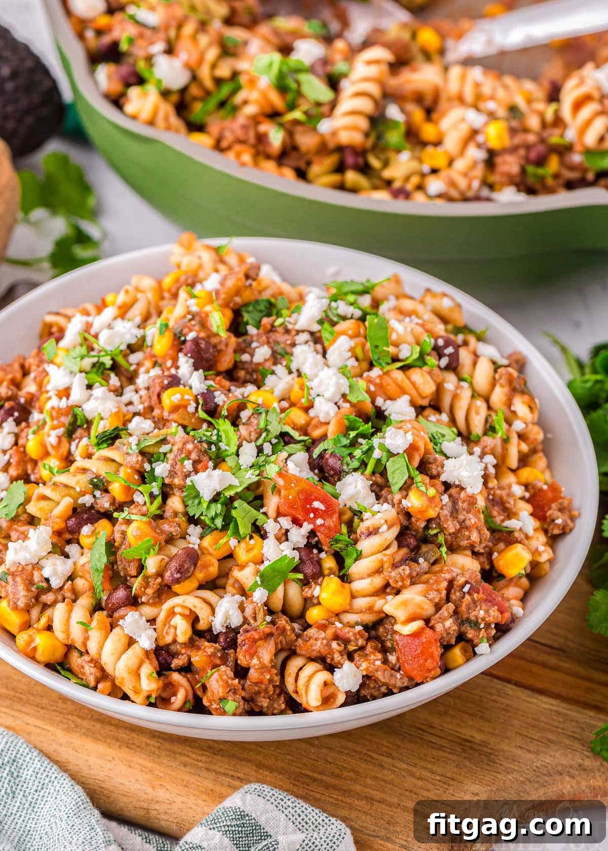 Angled view of a bowl filled with vibrant taco pasta, with a green skillet containing more pasta in the background.