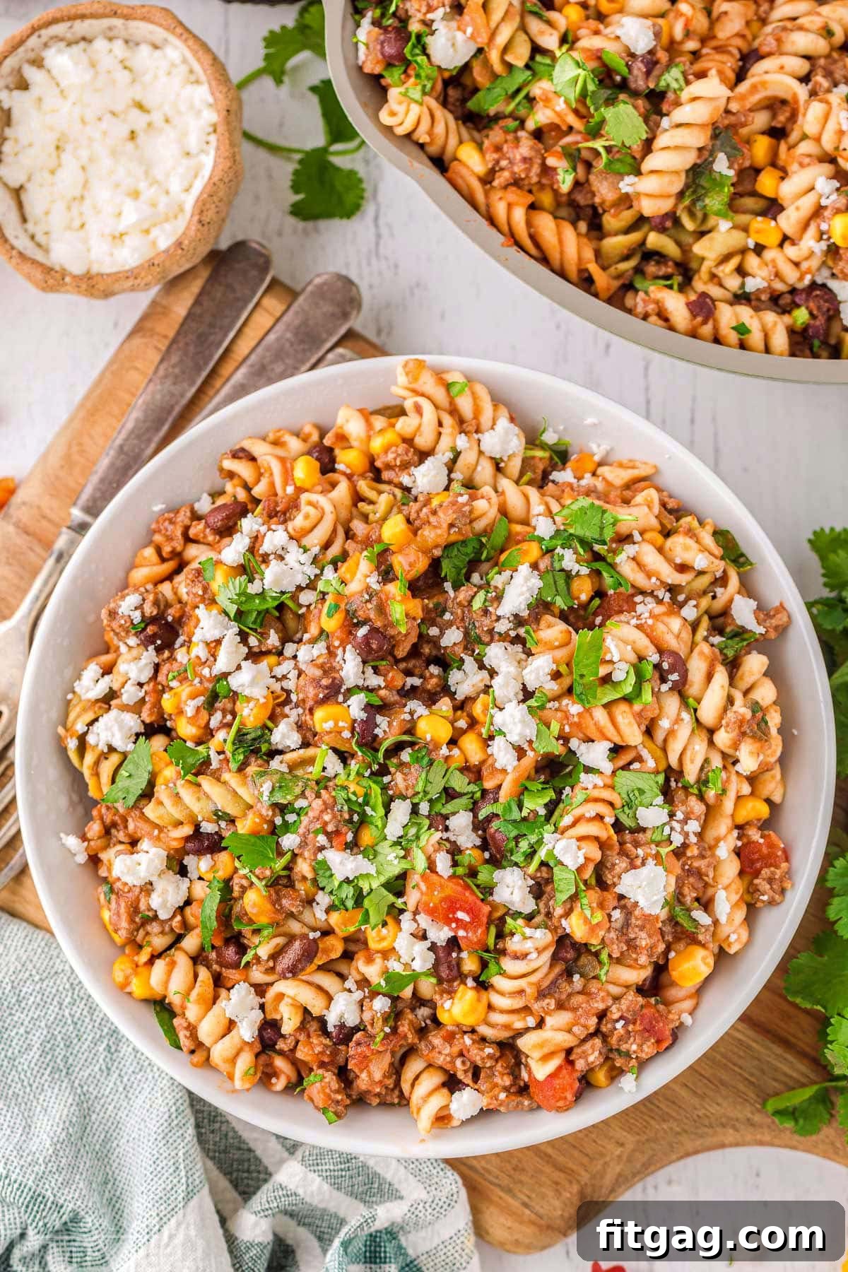 Top-down view of a serving of taco pasta in a round white bowl, with the green skillet containing the rest of the pasta visible in the background.