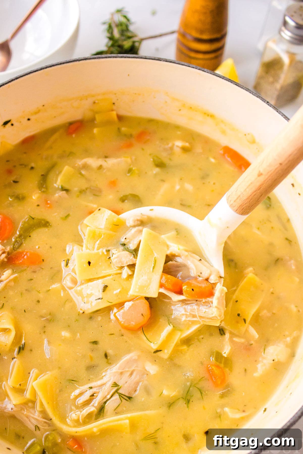 A serving spoon in a large pot of simmering creamy chicken noodle soup, showing tender chicken and noodles and steam rising.