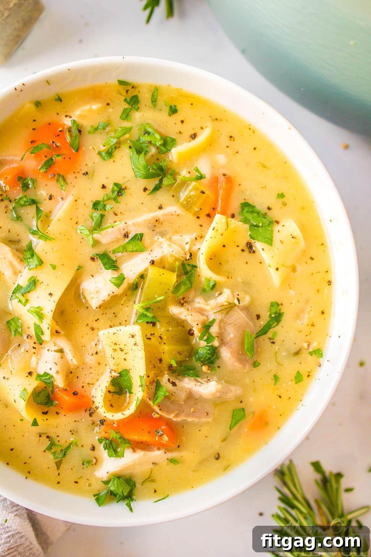 A top down close up shot of a white bowl of creamy chicken noodle soup, showing the rich broth, tender chicken, and perfectly cooked noodles, garnished with herbs.