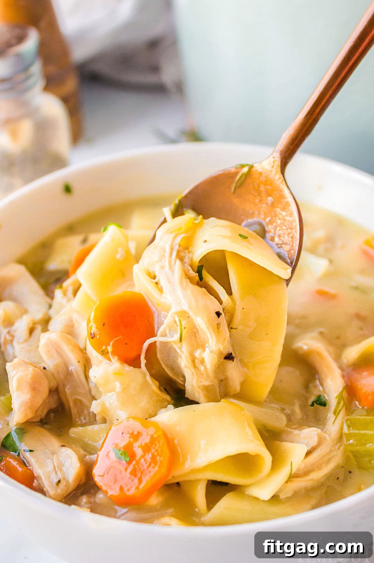 A spoon lifting tender chicken and noodles from a white bowl of creamy chicken noodle soup, garnished with fresh herbs, ready to be enjoyed.