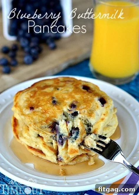 A stack of Blueberry Buttermilk Pancakes on a white plate with syrup drizzled over and a glass of orange juice in the background.