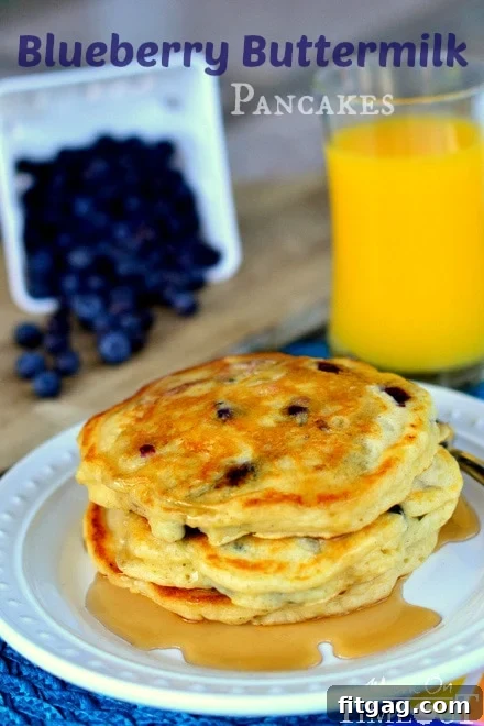A close-up of a generous stack of Blueberry Buttermilk Pancakes on a white plate with maple syrup, surrounded by fresh blueberries on a blue placemat.