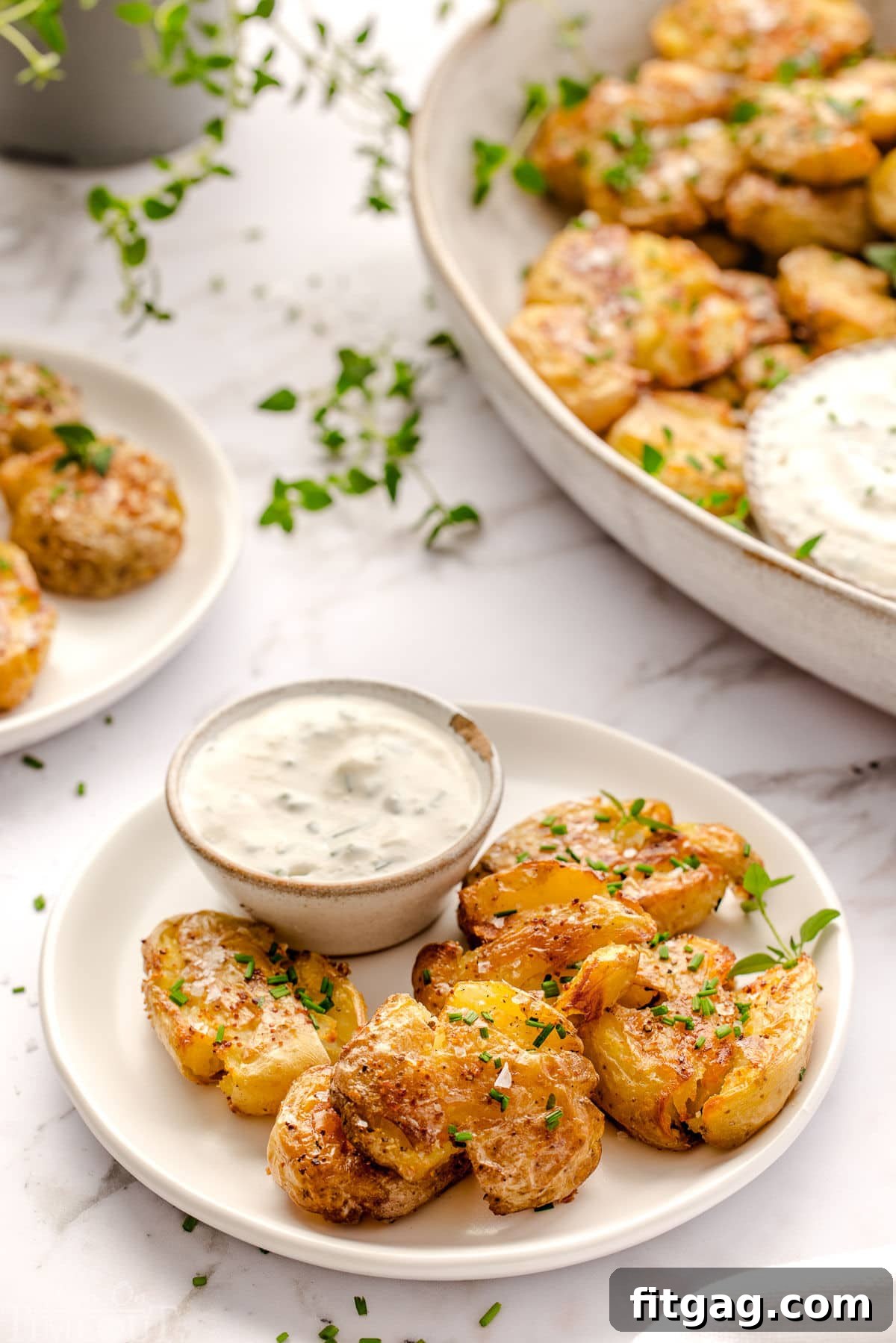 Smashed potatoes plated on a small round white plate with sour cream dip. Platter with more smashed potatoes in the background.