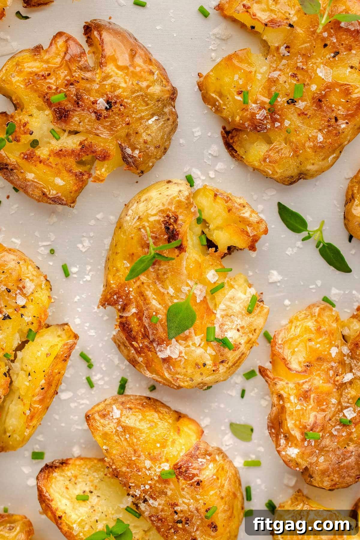 Top down view of smashed potatoes recipe on a white surface. The potatoes have been seasoned with salt, pepper and are topped with chives and thyme.