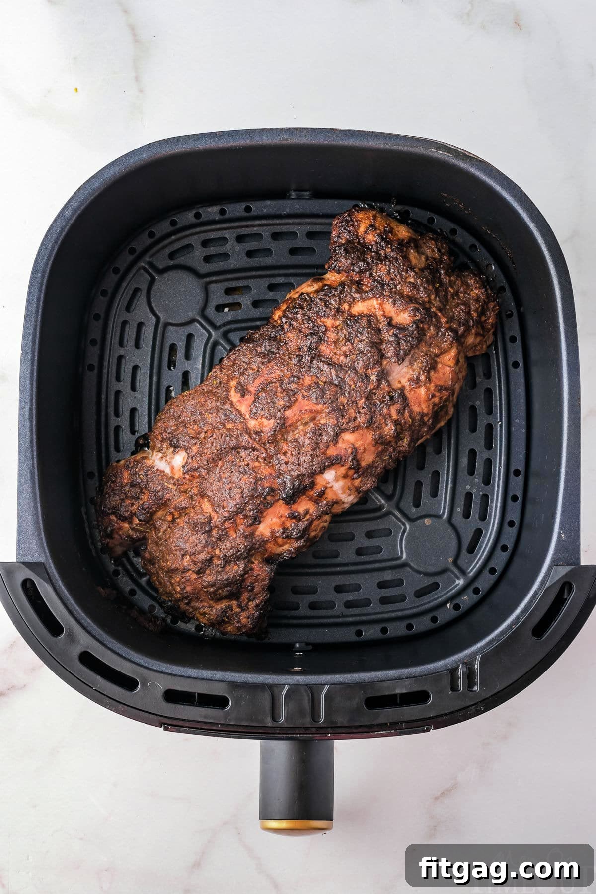Perfectly Tender Air Fryer Pork Tenderloin 3 Top-down view of a seasoned pork tenderloin cooking in an air fryer basket, sitting on a white surface.
