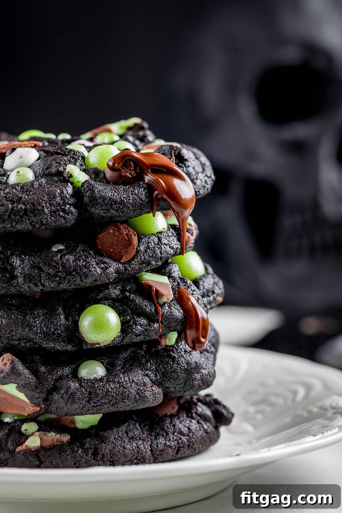 Side shot of stack of halloween cookies made with black cocoa powder stacked on a white plate with melty chocolate chips and green pearl candies.