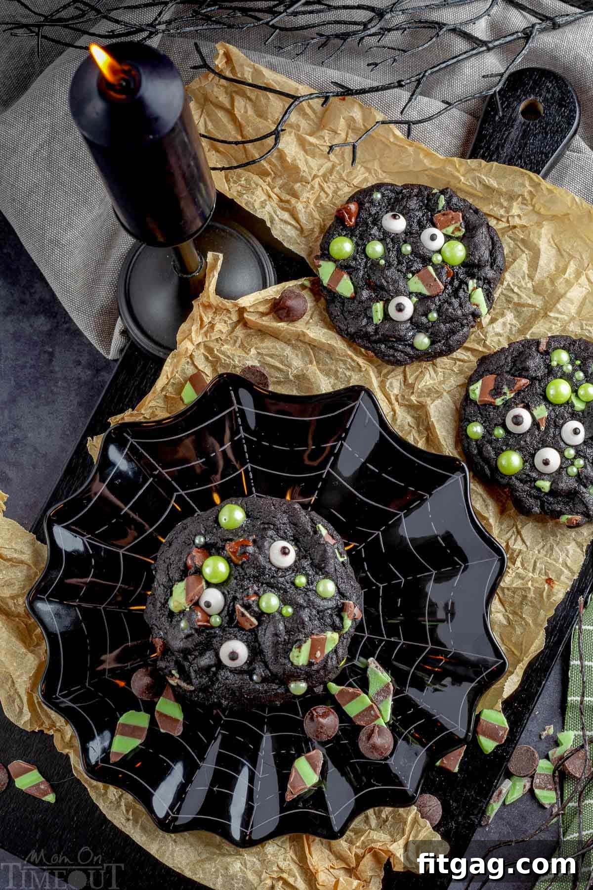 Halloween monster cookies on a black plate with a spider web design. Two cookies are on a piece of brown parchment paper. A black candle and black tree branch are next to them.