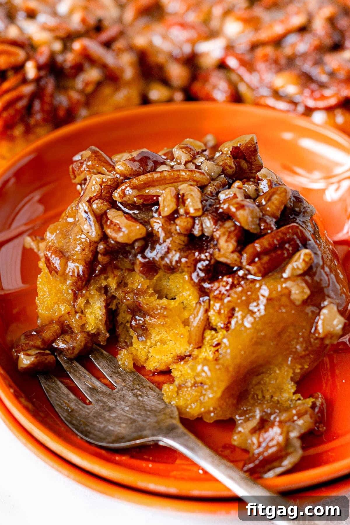 A single pumpkin sticky bun sits on an orange plate with a fork, a bite already taken, showcasing its soft interior and sticky caramel coating.