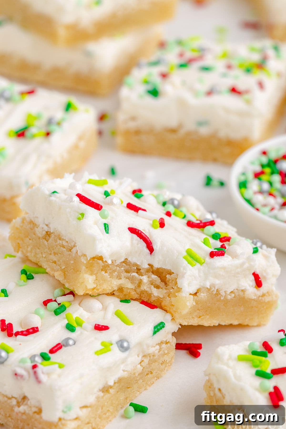 Festive Frosted Sugar Cookie Bars 9 A sugar cookie bar with a bite taken out, resting on another bar, with more bars in the background, emphasizing its deliciousness.