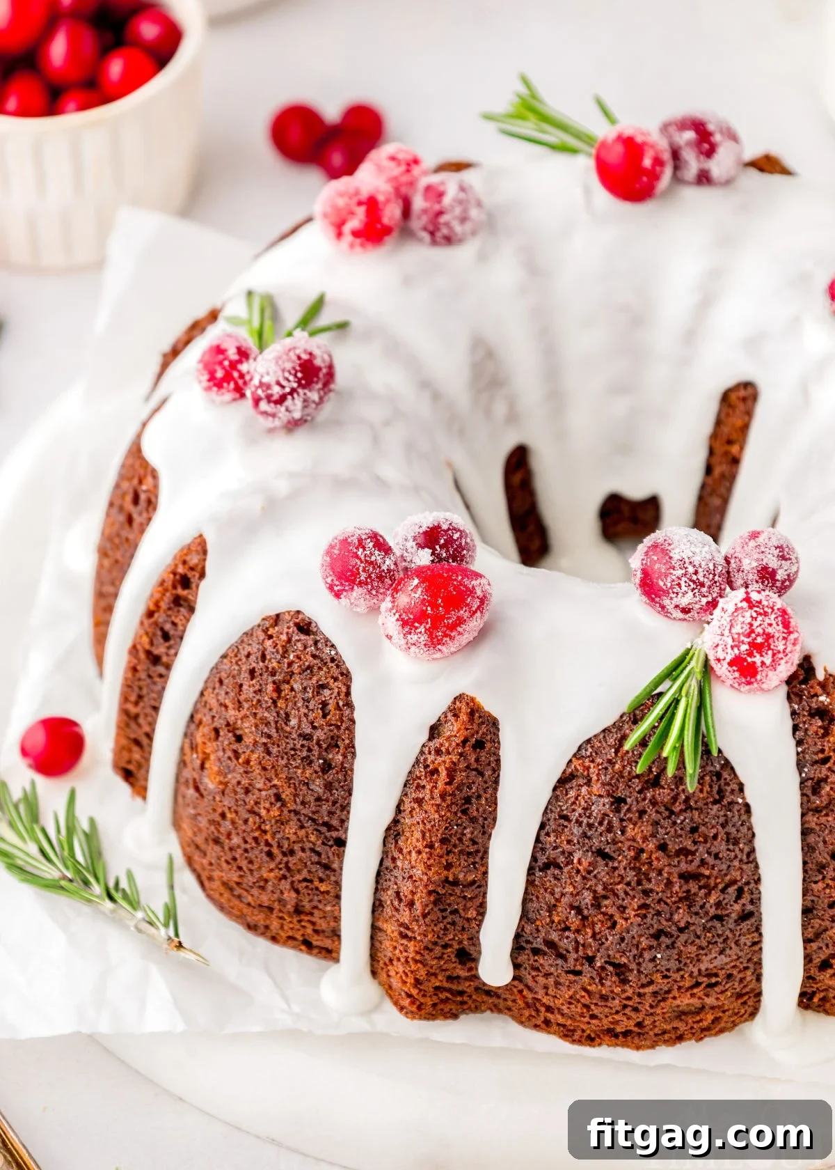 Gingerbread bundt cake topped with vanilla icing and cranberry and rosemary garnishes. The cake is on a white cake stand.