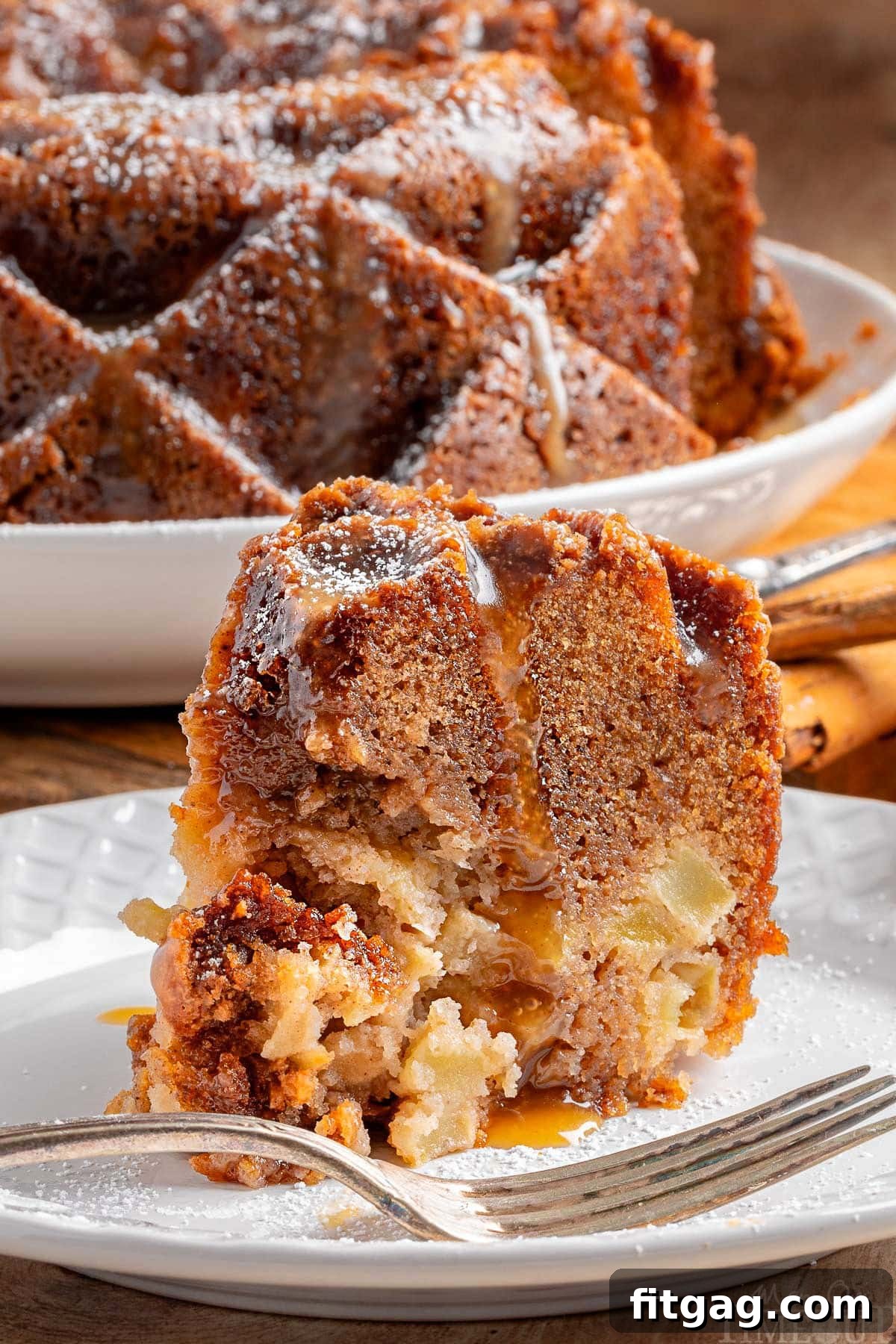 A beautiful slice of Caramel Apple Bundt Cake, generously drizzled with a rich bourbon caramel glaze, rests on a pristine white round plate. In the soft-focused background, the majestic full bundt cake waits, promising more deliciousness. This image perfectly captures the cake's moist texture and the glossy sheen of the glaze.