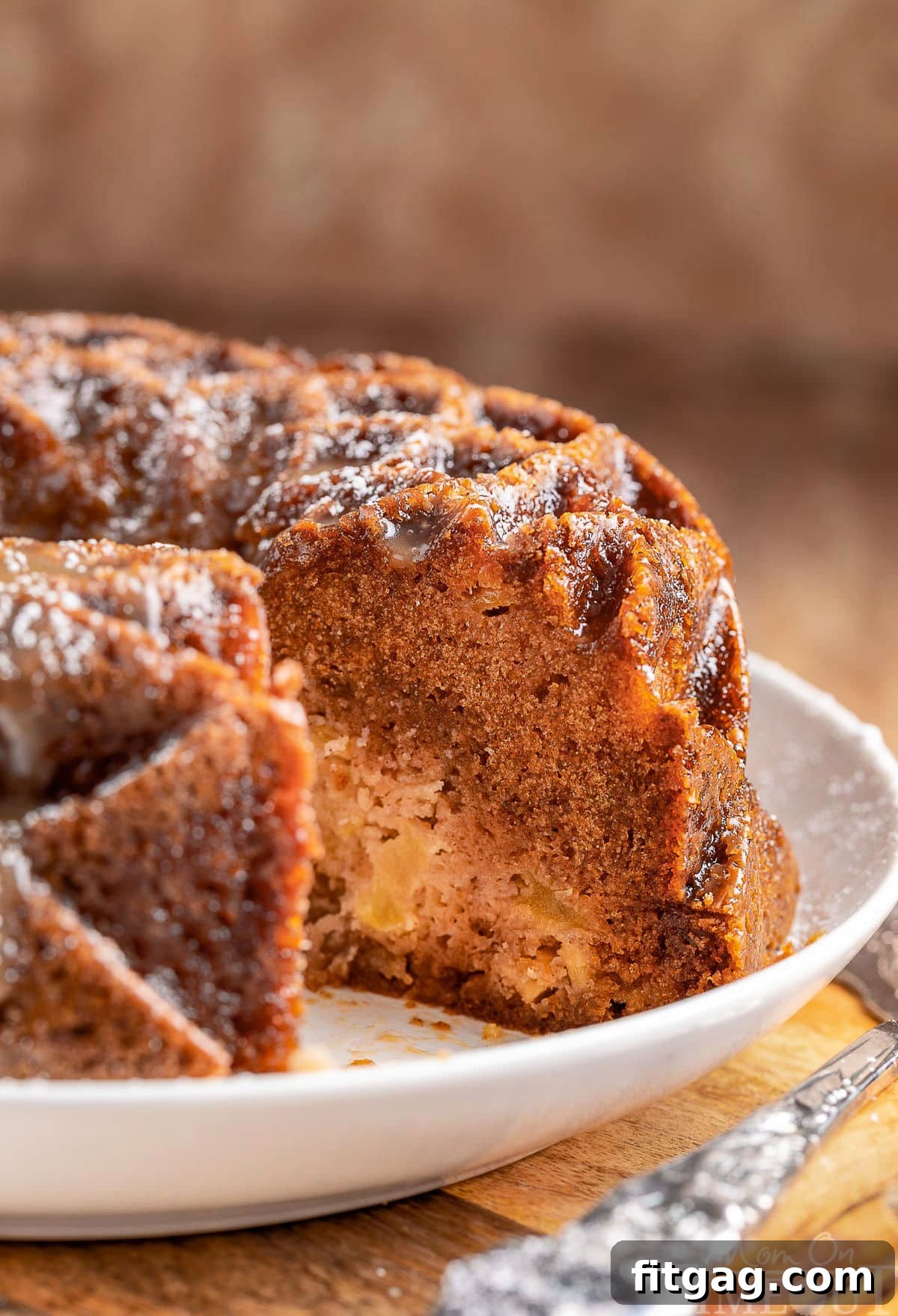 A perfectly baked Caramel Apple Bundt Cake, with a slice elegantly removed to reveal its moist, apple-flecked interior. The cake is presented on a pristine white serving dish, highlighting its golden-brown crust and inviting texture.