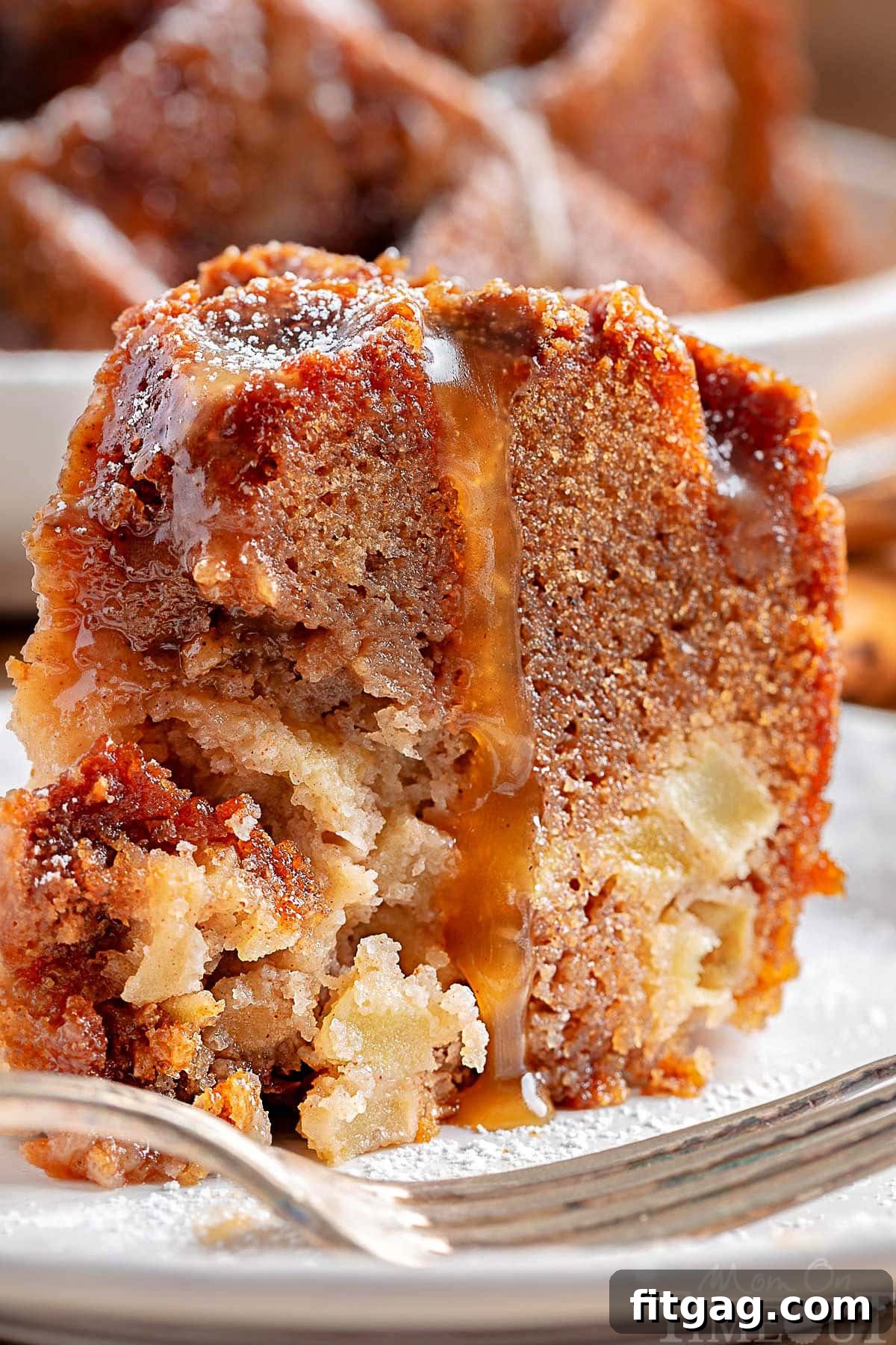 A close-up view of a single slice of Caramel Apple Bundt Cake, showcasing its moist, apple-filled interior and the generous coating of glistening caramel glaze. The slice sits on a white round plate, with the remainder of the beautifully glazed bundt cake subtly blurred in the background, creating an inviting focus on the texture and detail.