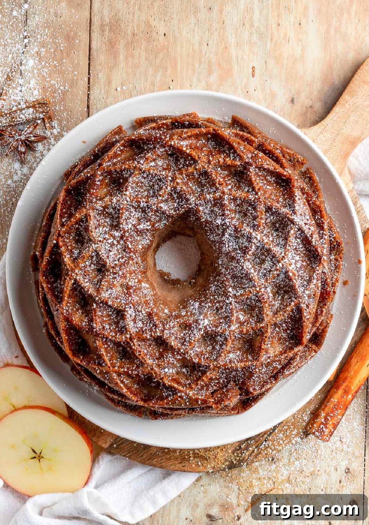 A beautiful Caramel Apple Bundt Cake, lightly dusted with a delicate layer of powdered sugar, presented on a pristine white serving plate. The elegant presentation highlights the cake's intricate bundt shape and suggests a soft, comforting sweetness.
