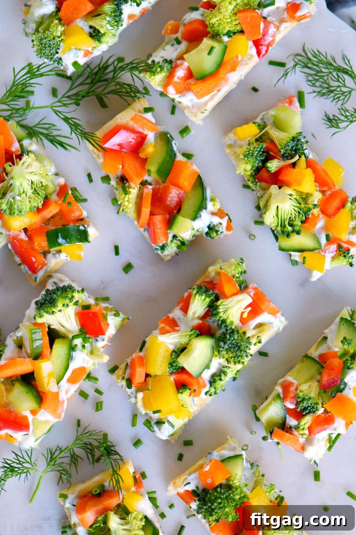 Party-Perfect Veggie Pizza Bites 3 A top-down view of freshly prepared veggie pizza, cut into small, serving-sized pieces and arranged on a marble cutting board, with scattered dill sprigs for garnish.