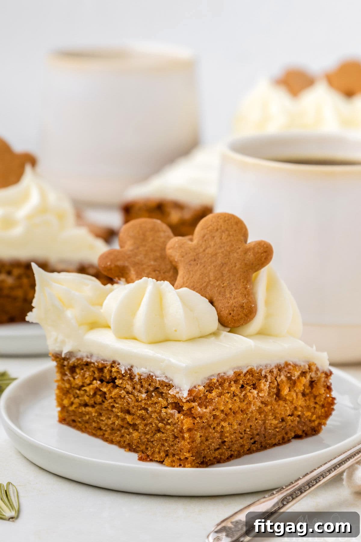 A slice of gingerbread cake topped with gingerbread men. The cake is on a white plate with mugs of coffee and more slices of cake in the background.