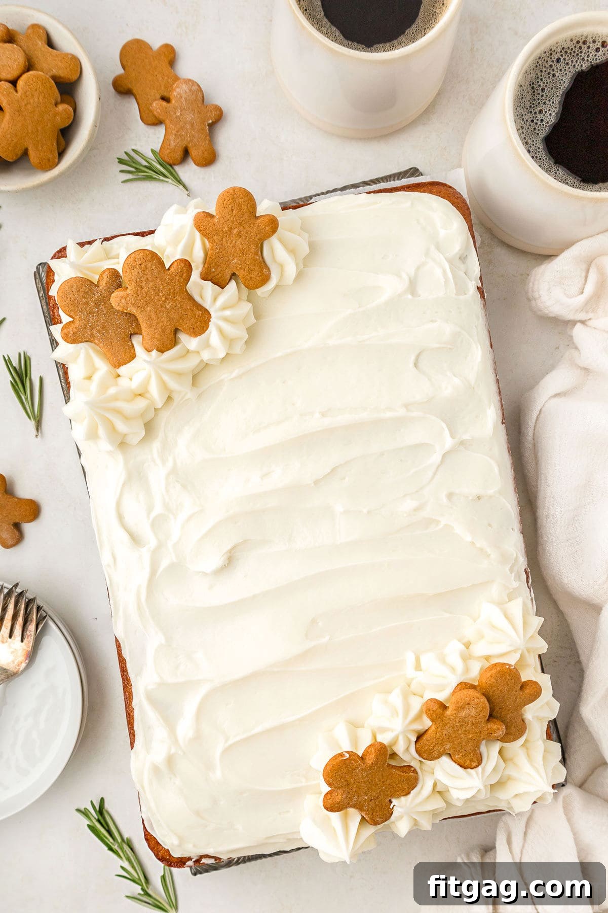 A top down view of gingerbread cake on a white surface. Mugs of coffee, gingerbread men, rosemary sprigs, plates, and a white linen are next to the ckae.