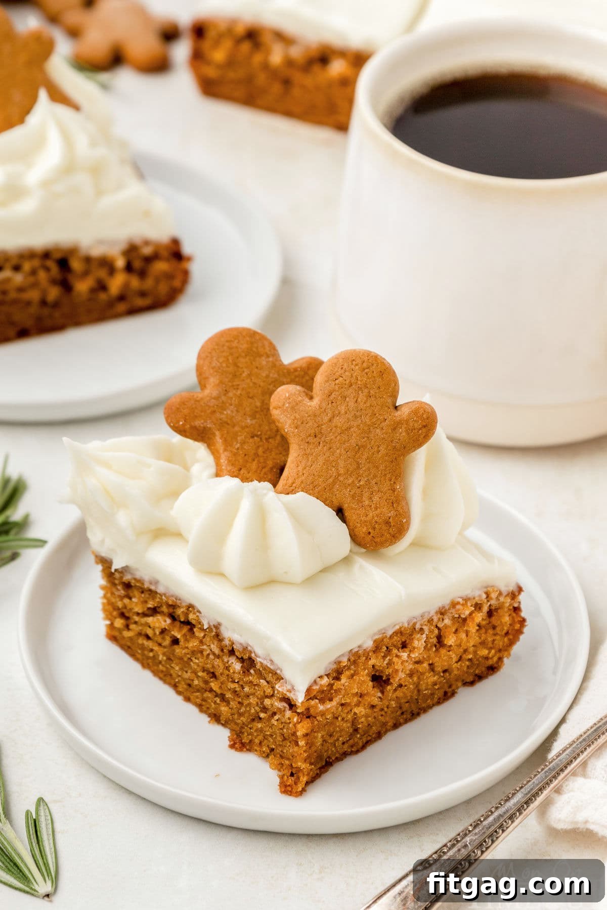 Angled view looking down on a piece of gingerbread cake topped with cream cheese frosting on a round white plate. A cup of coffee can be seen in the background.