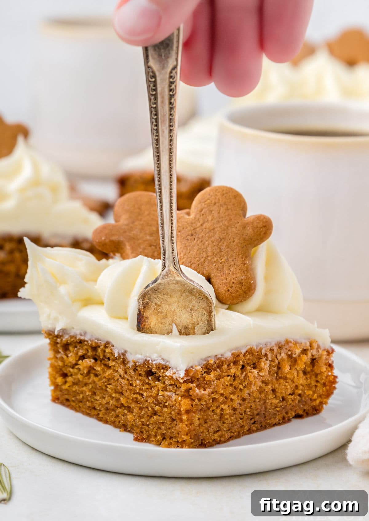 A hand putting a fork into a slice of gingerbread cake. The cake is on a white plate, and mugs of coffee and another piece of cake are in the background.