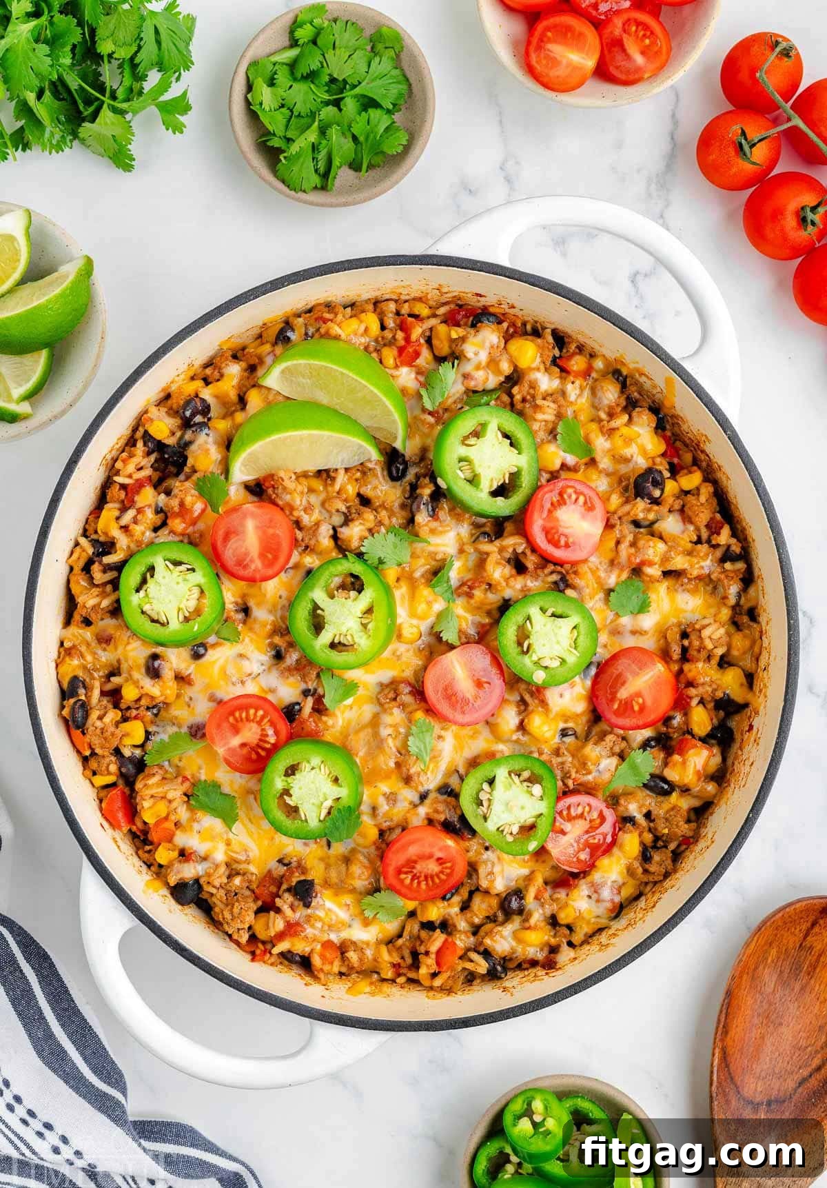 Top down view of taco rice in a white skillet topped with jalapeno slices, lime wedges and tomatoes. More taco topping can be seen scattered around the edge of the skillet.