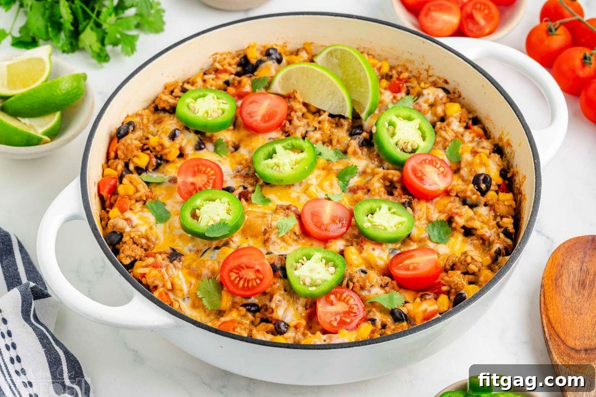 Top down angled view of taco rice in a white skillet topped with jalapeno slices, lime wedges and tomatoes. More taco topping can be seen scattered around the edge of the skillet.