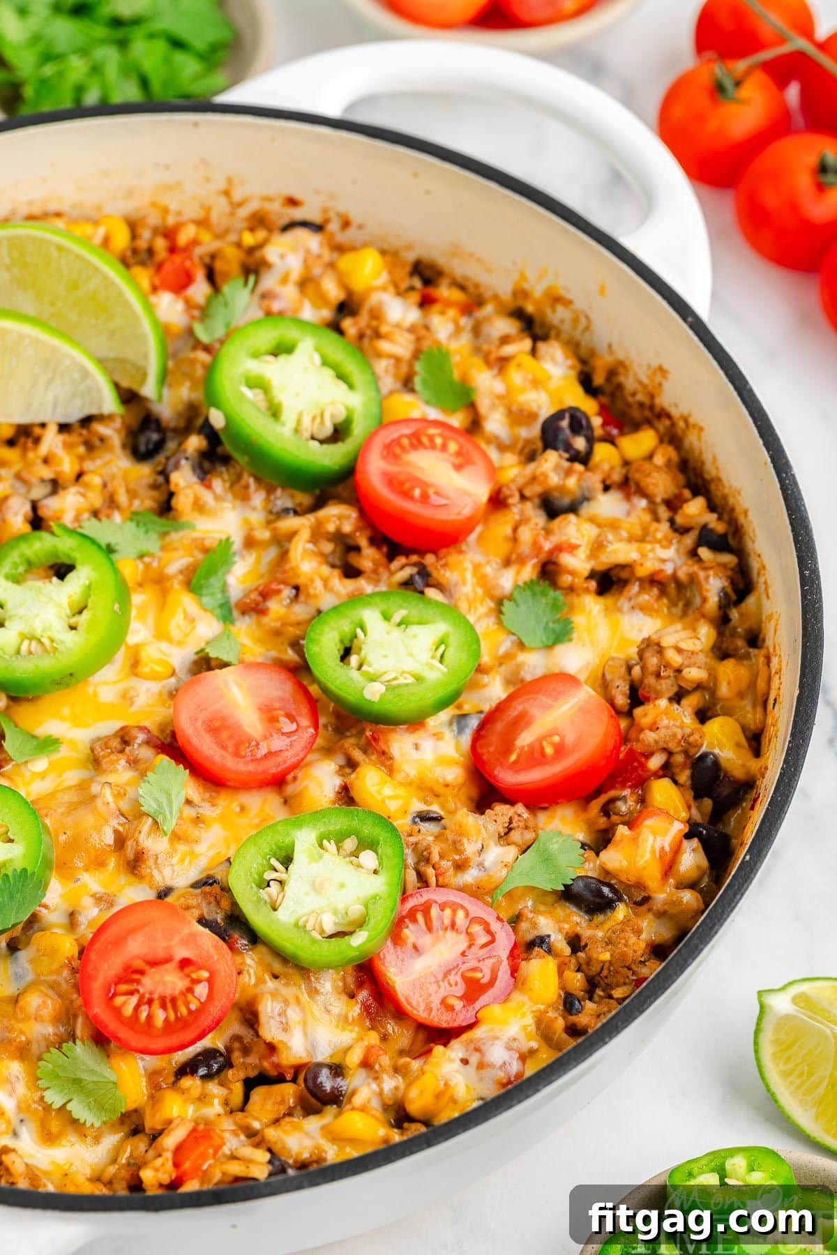 Close up view of cheesy taco rice in a white skillet, showcasing the melted cheese and vibrant ingredients, garnished with fresh jalapeno slices, lime wedges, and diced tomatoes around the rim.