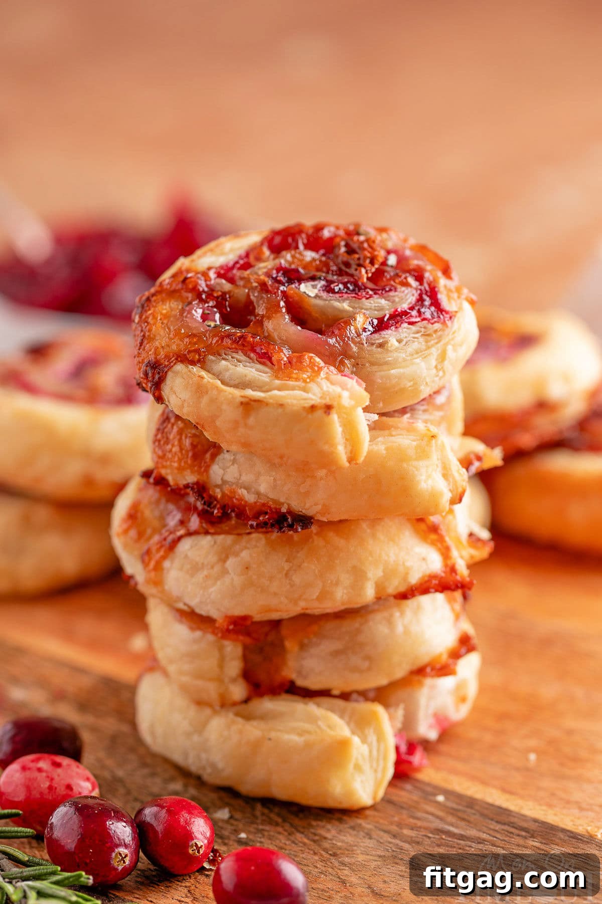 Elegant Cranberry Brie Spirals 3 Five cranberry brie puff pastry appetizers stacked elegantly on a rustic wood board, with fresh cranberries and rosemary sprigs scattered around.