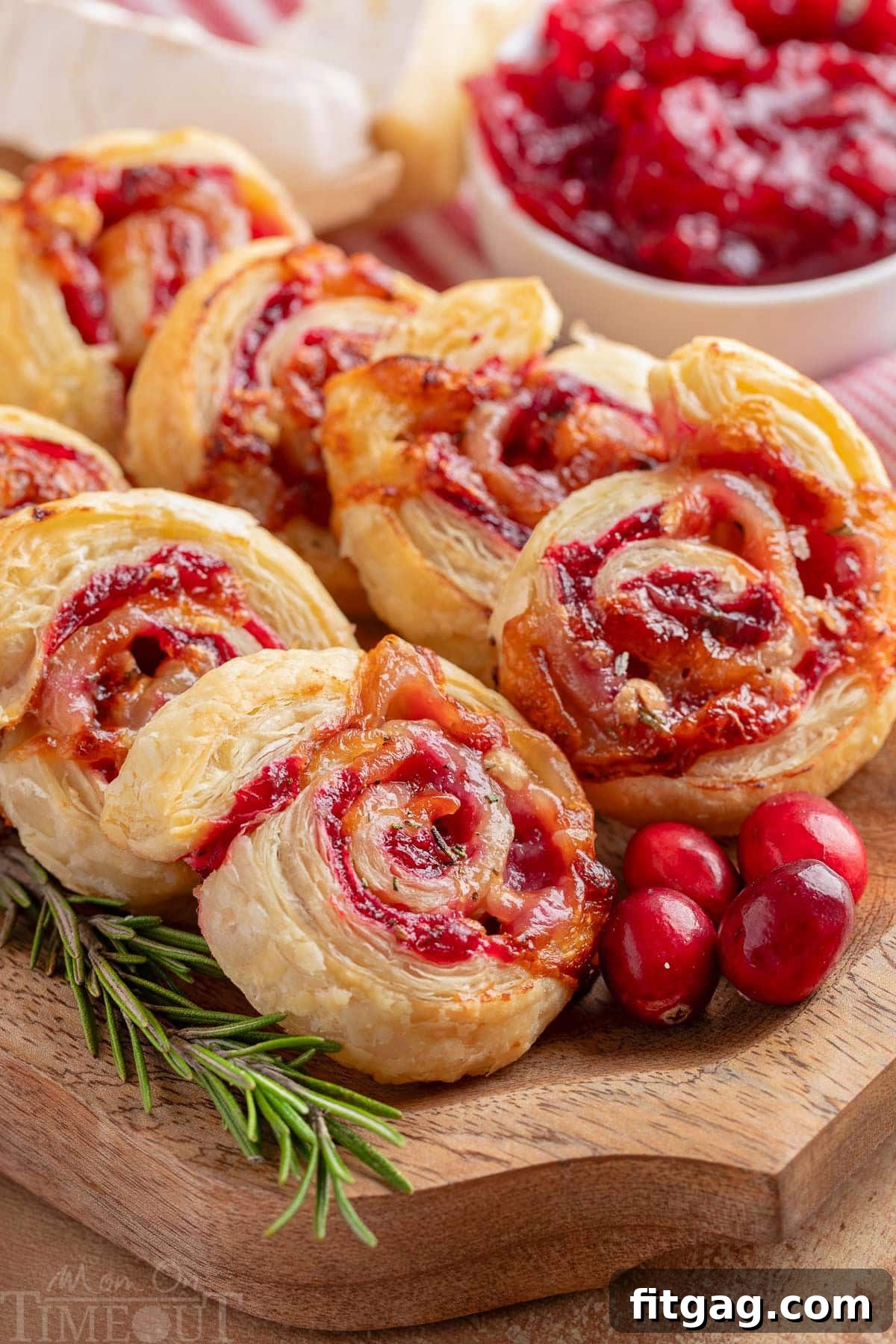 Elegant Cranberry Brie Spirals 6 Wood serving tray filled with freshly baked cranberry brie pinwheels, adorned with fresh rosemary and scattered cranberries for a festive display.