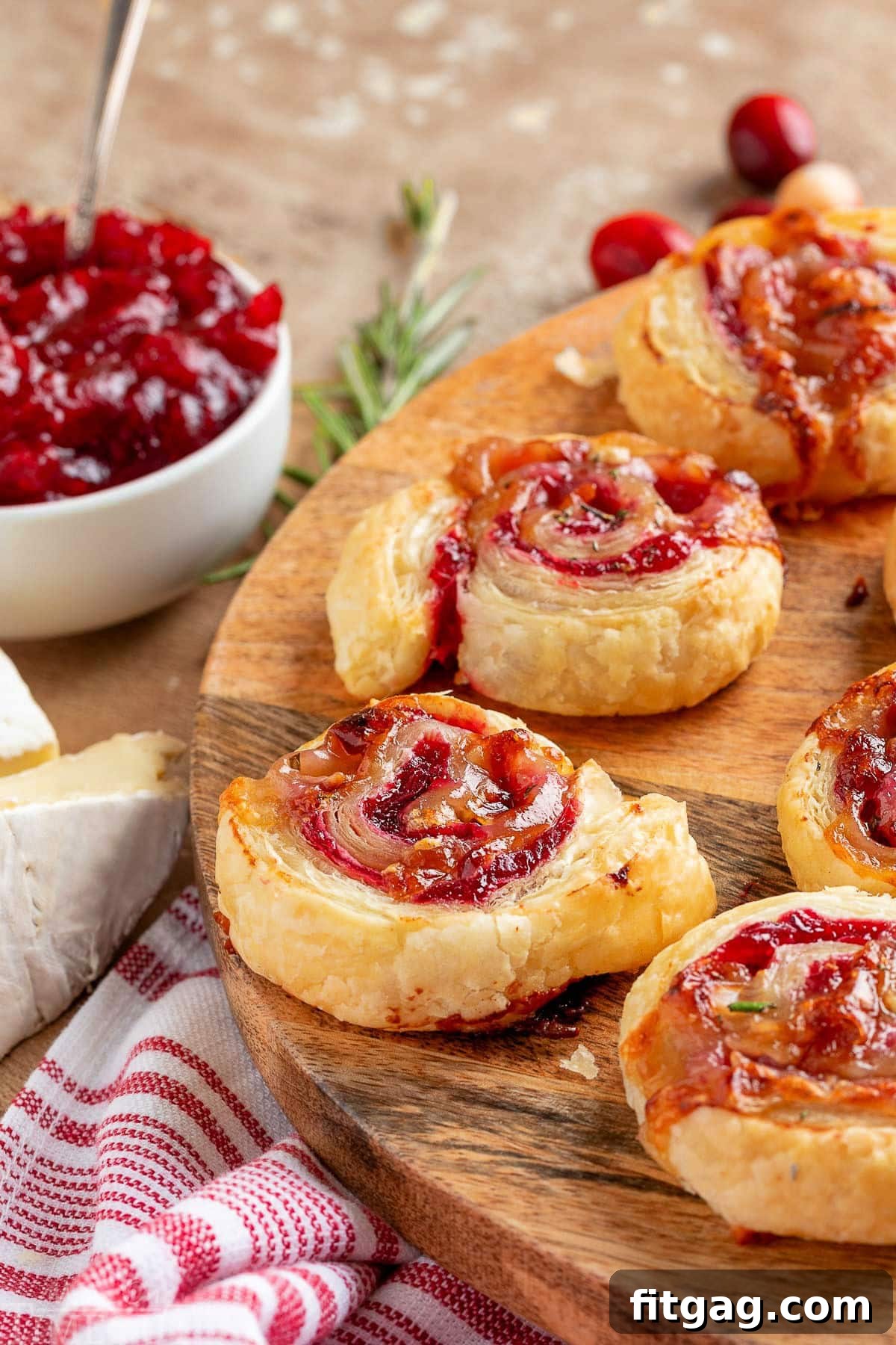 Elegant Cranberry Brie Spirals 8 Cranberry brie puff pastry pinwheels artfully arranged on a rustic wood serving board, with a bowl of cranberry sauce in the soft-focus background.