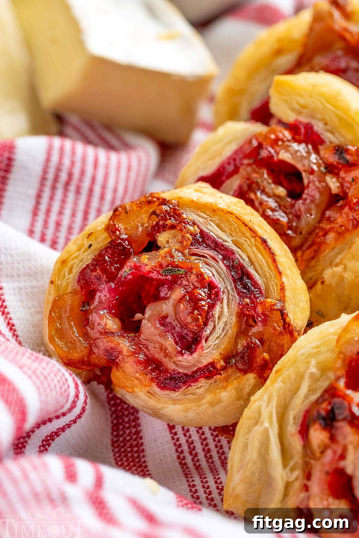 Elegant Cranberry Brie Spirals 9 Cranberry brie appetizers made with puff pastry, elegantly presented in a charming basket lined with a festive red and white striped towel, highlighting their holiday appeal.