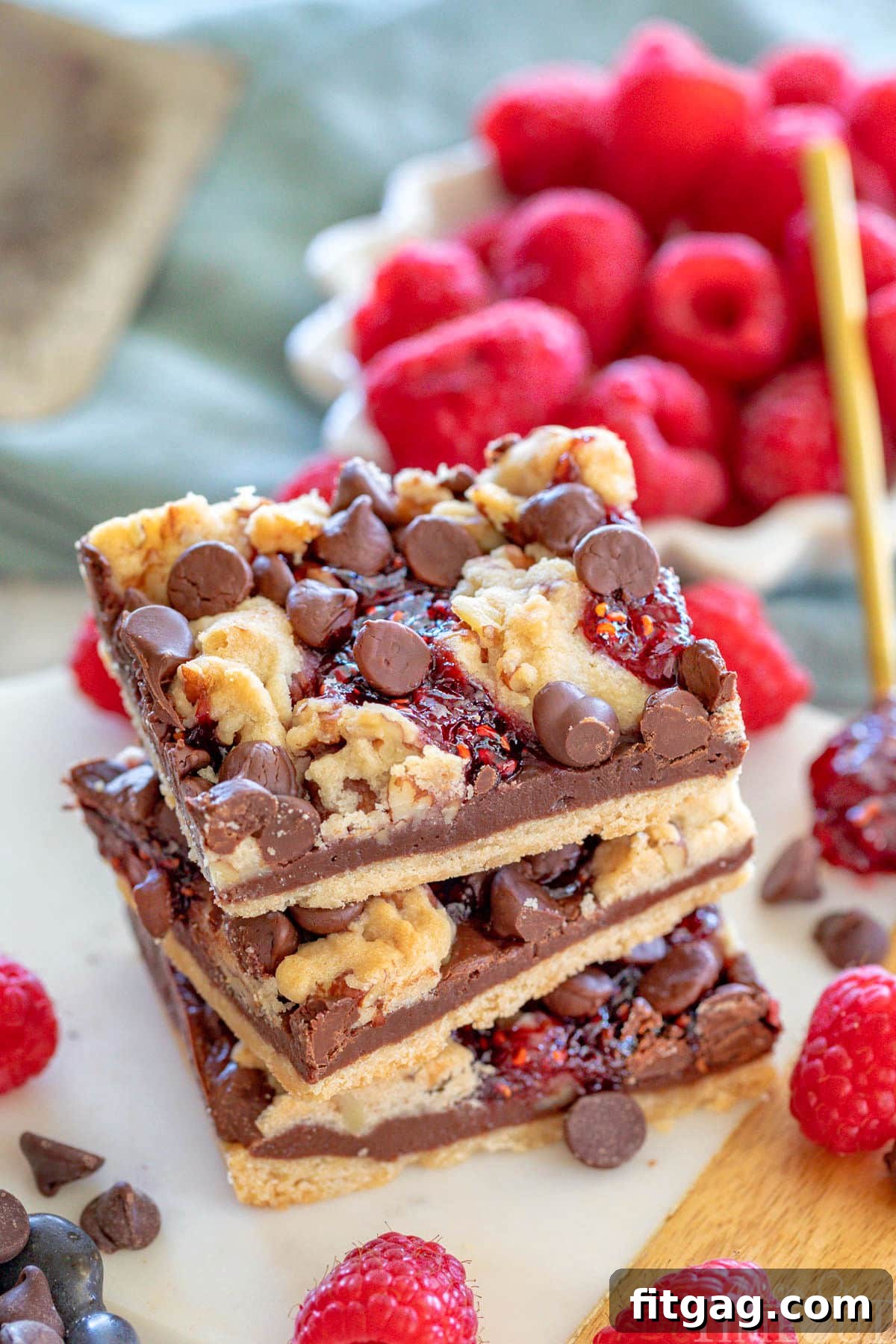 Decadent Raspberry Crumble Squares 2 Three chocolate raspberry crumb bars stacked on a white marble board. Fresh raspberries can be seen in a white bowl in the background.