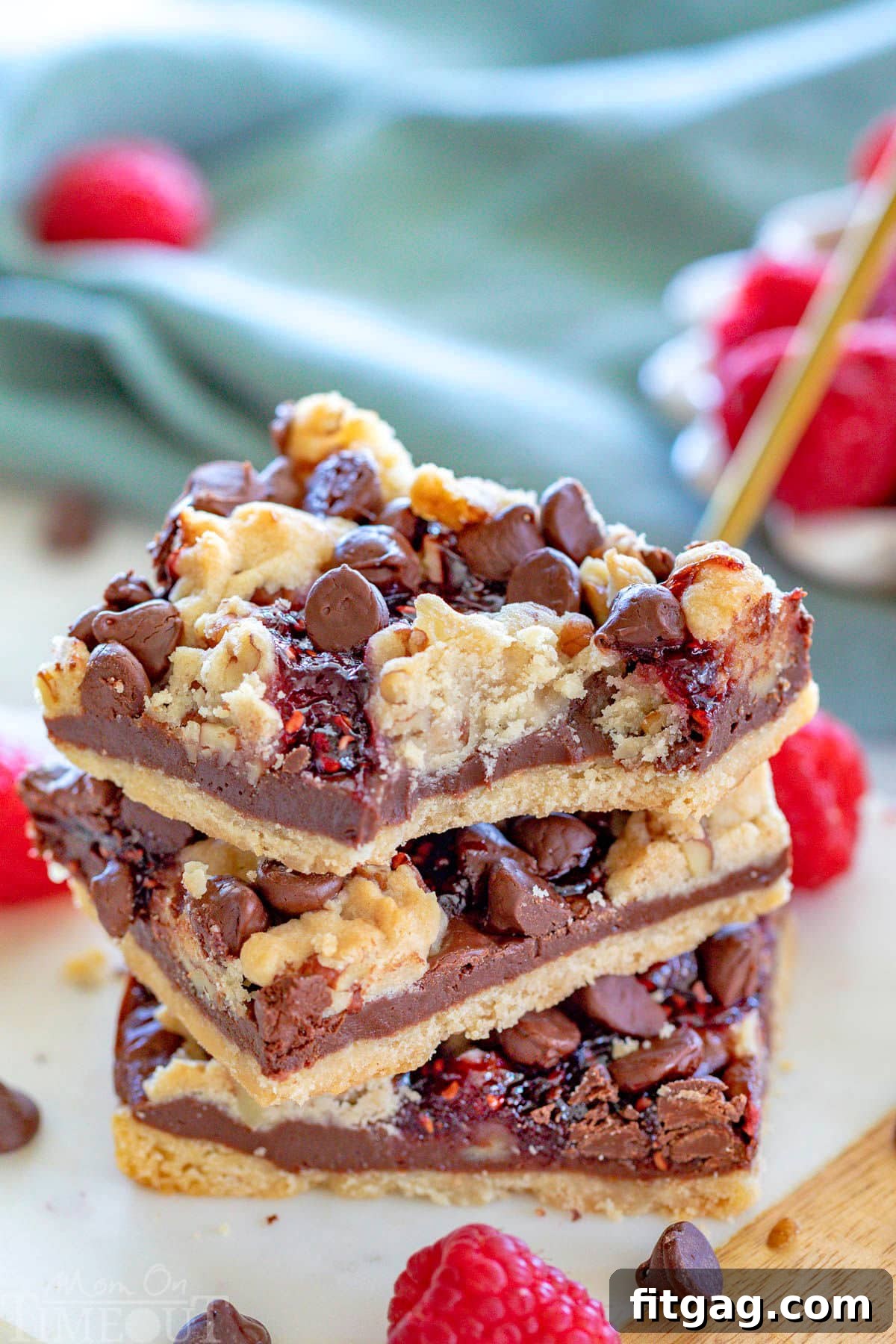 Decadent Raspberry Crumble Squares 4 Three chocolate raspberry crumb bars stacked on a white marble board. Fresh raspberries can be seen in a white bowl in the background. The top bar has a bite taken from it.