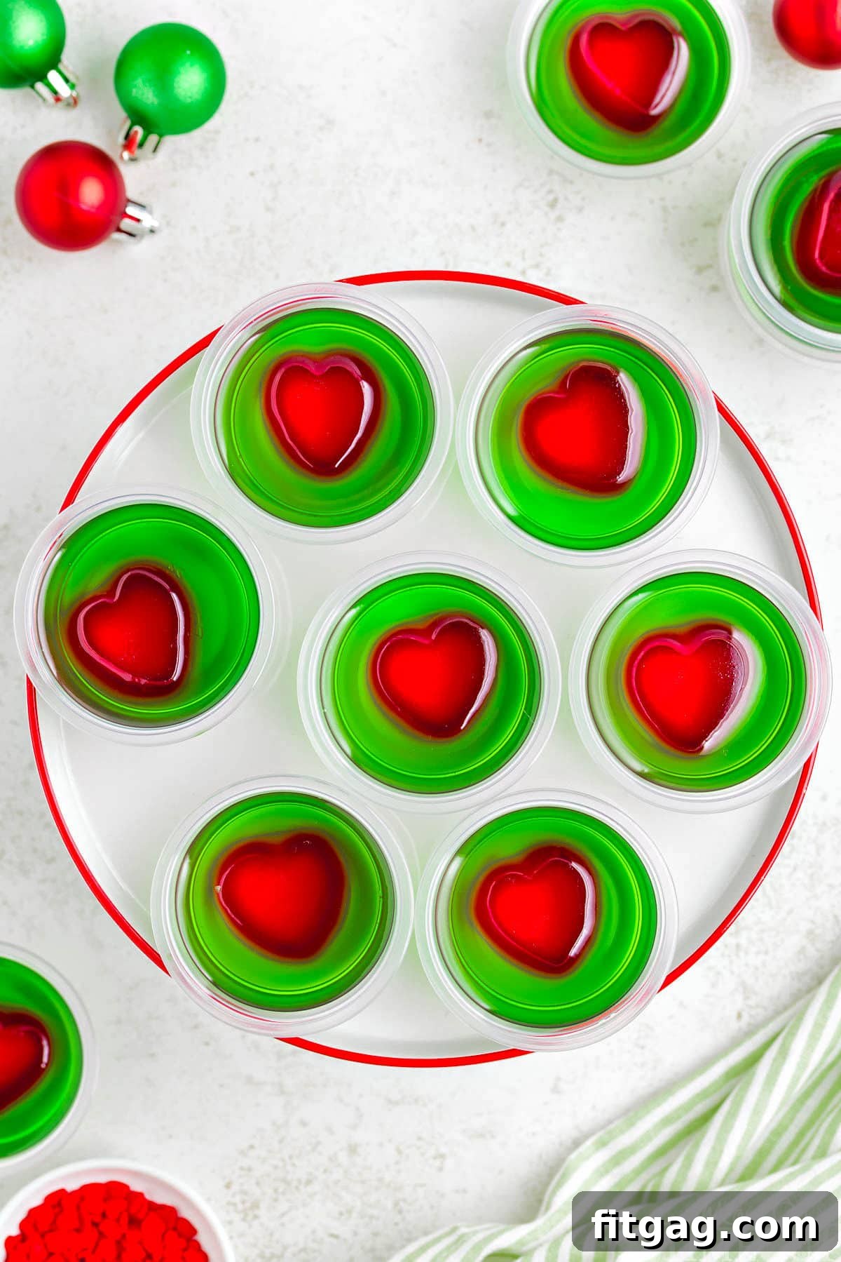 A top down view of grinch jello shots on a white plate with a red rim. Each shot is green with a red heart in the center.