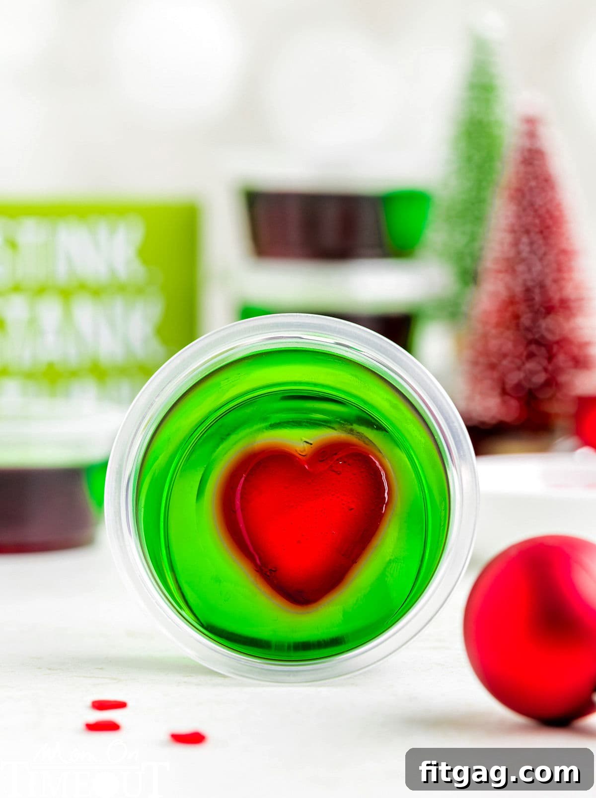 A green and red grinch jello shot in a shot glass laying on its side with Christmas decorations in the background.